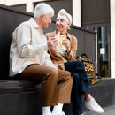Two seniors sitting and smiling outdoors, holding sandwiches, with a leopard print Inogen carry bag beside them.