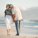 Older couple holding hands walking on beach, woman wearing portable oxygen backpack carrier.